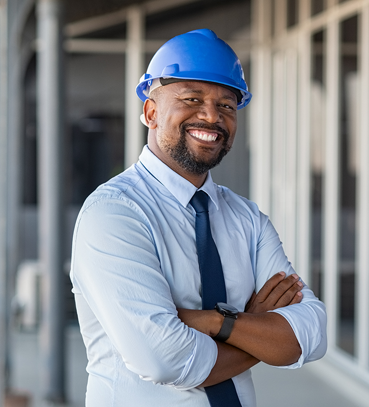 Portrait of african american man architect at building site with folded arms looking at camera. Confident construction manager in formal clothing wearing blue hardhat. Successful mature civil engineer at construction site with copy space.